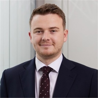 Matthew Lilley in a black jacket and brown spotted tie, in front of a pale background