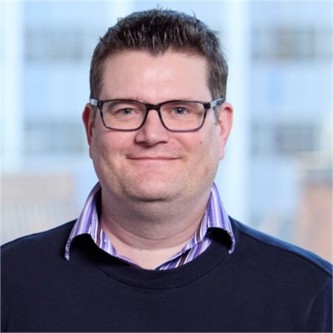 James Wood in glasses, a navy jumper and purple striped shirt, in front of a window