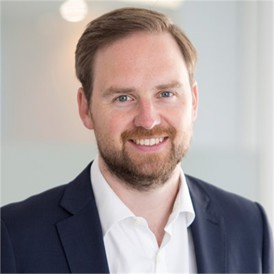 Tim Whitney wearing a white formal shirt, navy blue blazer in front of a blurry background.