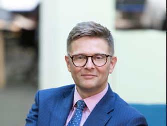 Andrew Burwood wearing a pink shirt, blue tie and a blue blazer with a blurry office background