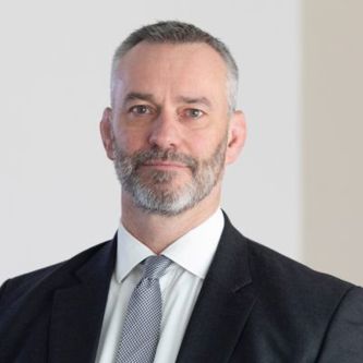 Richard Bedfore wearing white formal shirt, light grey tie, dark grey suit with a blurry background of a wall.