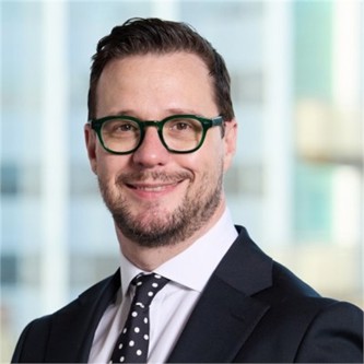 Jody Kite in glasses, a black jacket and a black spotty tie, in front of a window