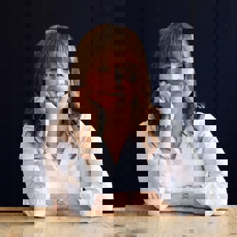 Sarah Petherbridge wearing a white shirt with her elbow propped on a table and chin leaning on hand. 