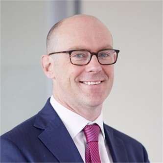 Martin Priestley in glasses, a black jacket and pink tie, in front of a pale background