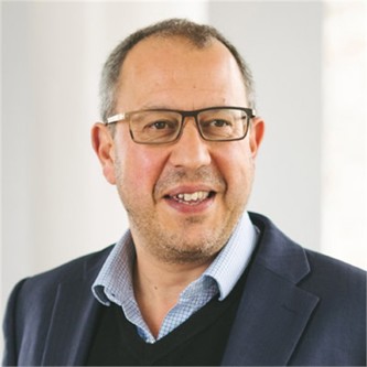 Nick Stone in glasses, a dark jacket, black jumper and checked blue shirt, in front of a pale background