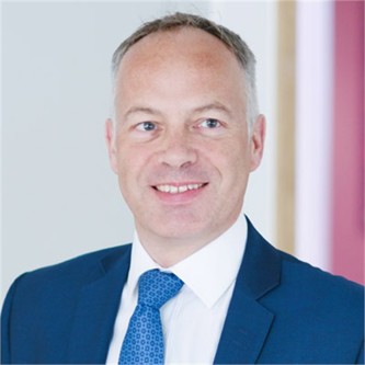 Greg Gibson in a blue suit and tie, in front of a pale background
