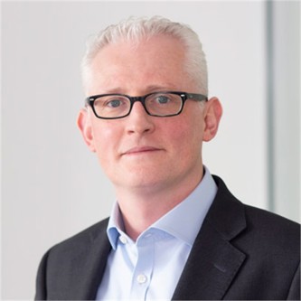 Neil Howes in glasses, a dark jacket and pale blue shirt, in front of a grey background