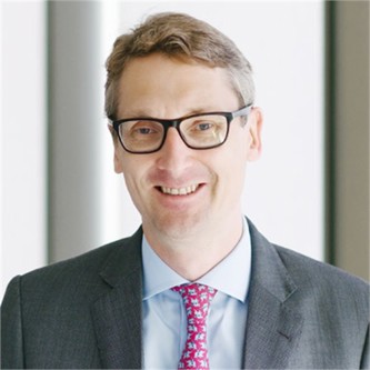 Tom Pickthorn wearing glasses, pink tie, light blue formal shirt, grey blazer in front of a blurry background.
